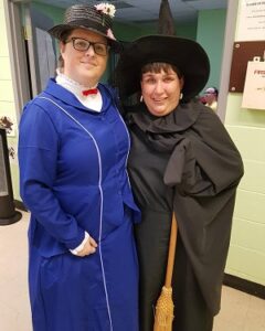 Two women standing beside each other one is wearing a blue dress and hat and the other is wearing a black dress, a witch’s hat and is holding a broom. 