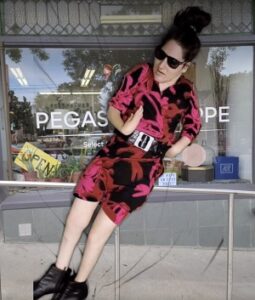 A woman wearing a black and pink dress with sunglasses and black boots is in front of a store window.