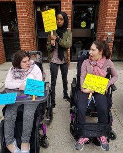 Two women in wheelchairs have signs in front of them while another woman is standing behind them with a yellow sign. 