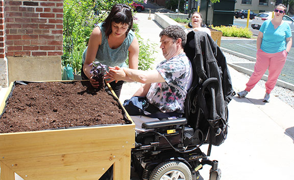 A Pegasus participant in a wheelchair assisting a volunteer in front of a wooden box to plant seeds in.