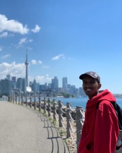 A man wearing a red sweater and hat standing by the water the CN tower can be seen in the distance.