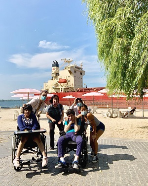 A group of people huddled around on a boardwalk there is sand and water in the background.