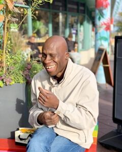 A man in a beige sweater sitting by a planter with plants.