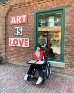 A woman in a wheelchair is in front of a brick wall with the words "Art Is Love" in the background.