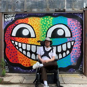 A man is sitting in front of a rainbow mural on a brick wall.