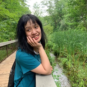 A woman in a green shirt is standing with her hand on railing overlooking a stream.