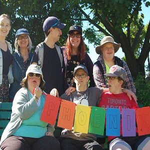 A group of Pegasus participants and staff holding a multi-coloured banner that says "Phoenix". The group is outside at the annual picnic.