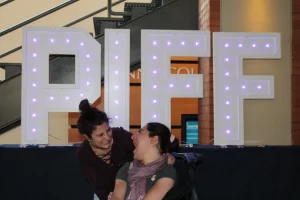 A picture of a Pegasus participant and a support worker smiling at each other in front of a sign that says "PIFF"