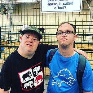 Two men standing with their arm around each other one is wearing a blue shirt and the other is wearing a black shirt and hat.