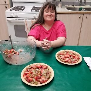 A woman in a pink shirt sitting in front of a green table with two pies and a bowl of strawberries beside her.