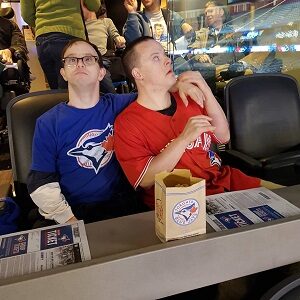 Two men sitting beside each other one is wearing a Blue Jays shirt and has his arm around the man in a red Blue Jays jersey there is popcorn in front of them.