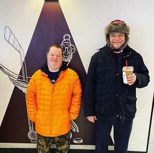 A man wearing a yellow puffer coat is standing next to another man wearing a black coat and winter hat who is holding a drink.