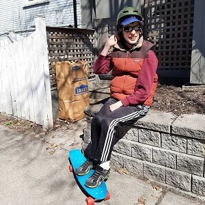 A man wearing a helmet and vest is sitting on a stone wall with his feet on a blue skateboard.