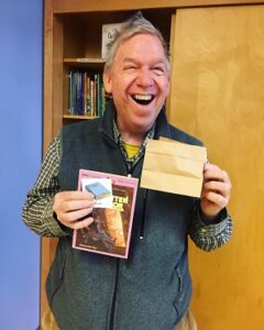 A man wearing a grey vest and plaid shirt holding a book and a brown bag.