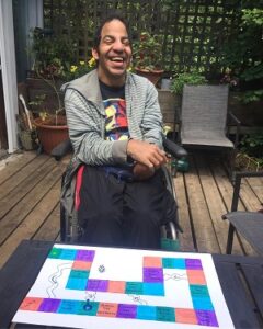 A man in a wheelchair wearing a grey striped sweater is on a deck with a homemade board game in front of him.