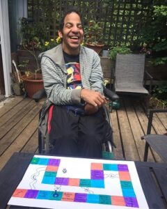A man in a wheelchair wearing a grey striped sweater is on a deck with a homemade board game in front of him.