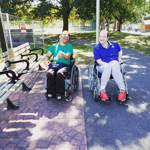 Two men in wheelchairs are beside each other one is wearing a green shirt and black pants the other is wearing a blue shirt and grey pants they are at a park.