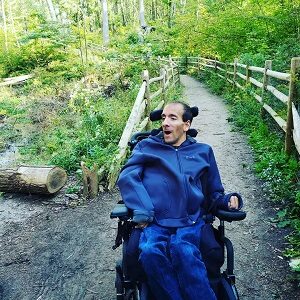 A man in a wheelchair wearing blue is on a path in a forest.