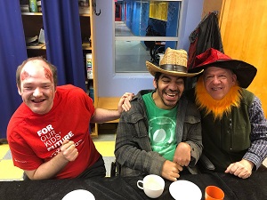 Three men are sitting beside each other at a table one is wearing a red shirt, the man in the middle is wearing a green shirt and a hat and the other man is wearing a hat, a yellow beard, and a green vest.